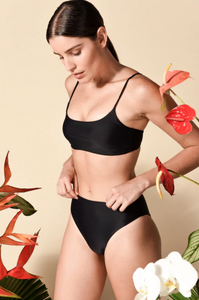 Model standing against wall among foliage while wearing the Hinge Top and Incline Bottom in black
